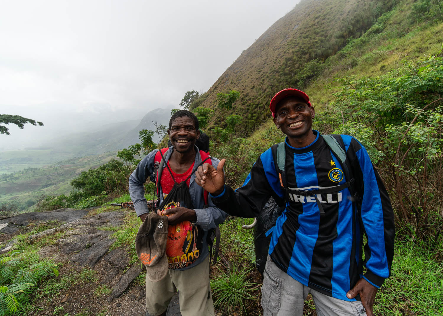 Two hikers on a mountain trail with a scenic view of foggy mountains and greenery.