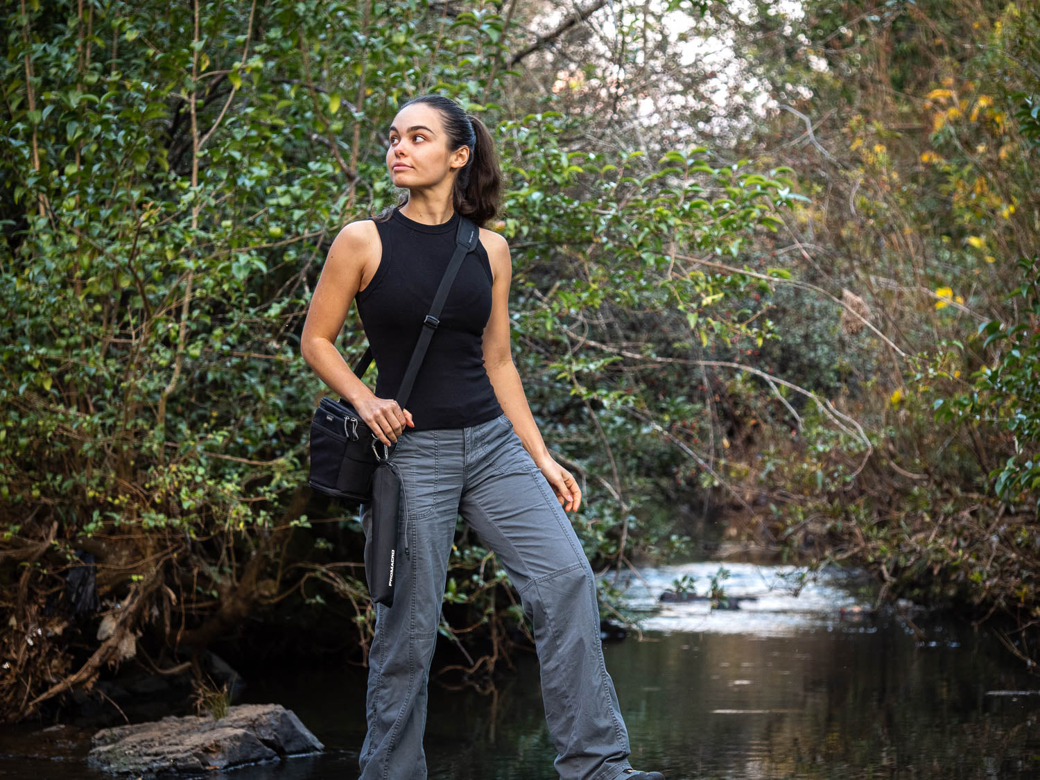 Woman standing by a stream in a forested area, wearing a black tank top and gray pants and promacro diffuser pouch